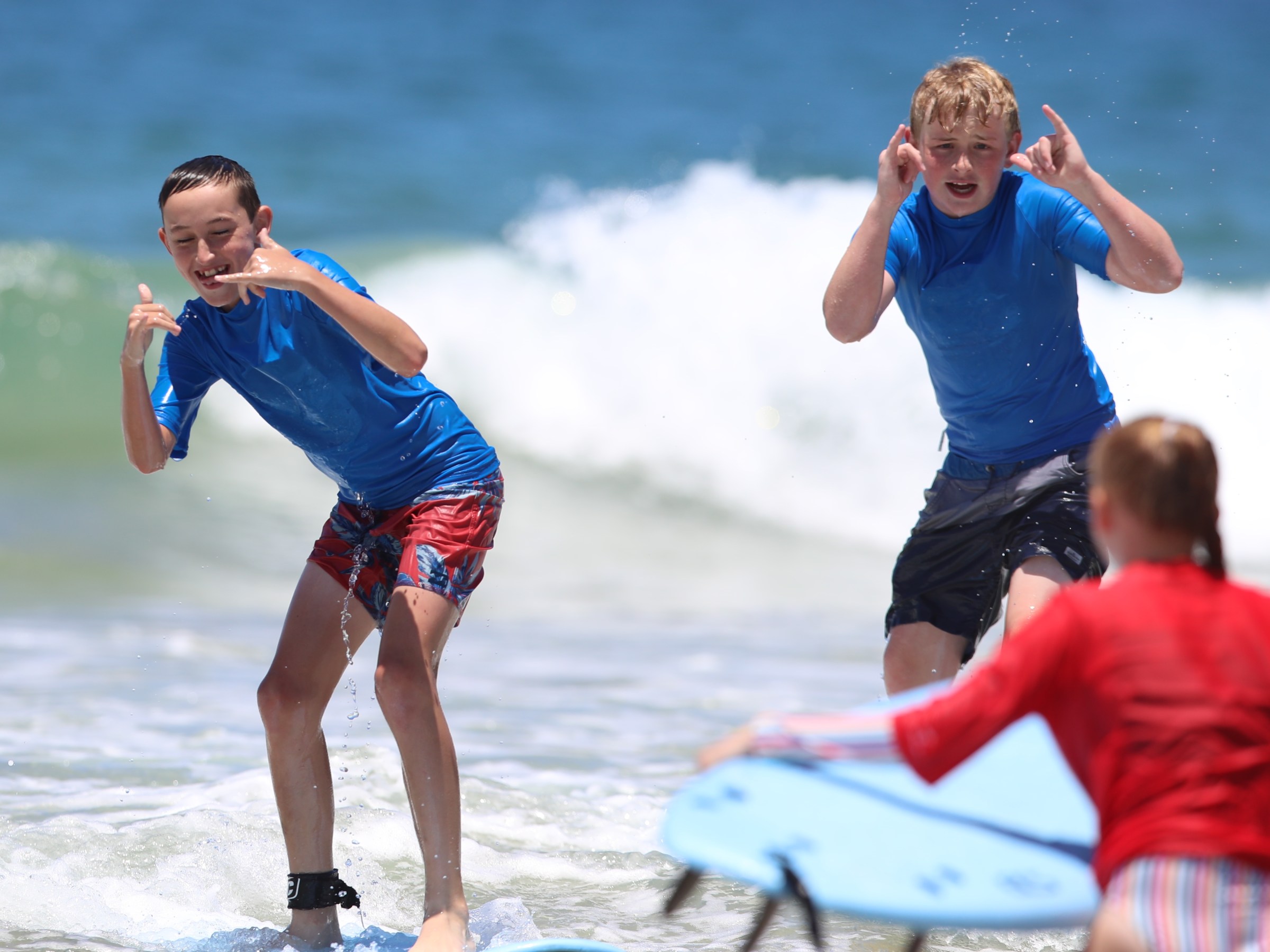 Two boys in blue shirts playing in the ocean waves, a girl with a surfboard in the foreground.